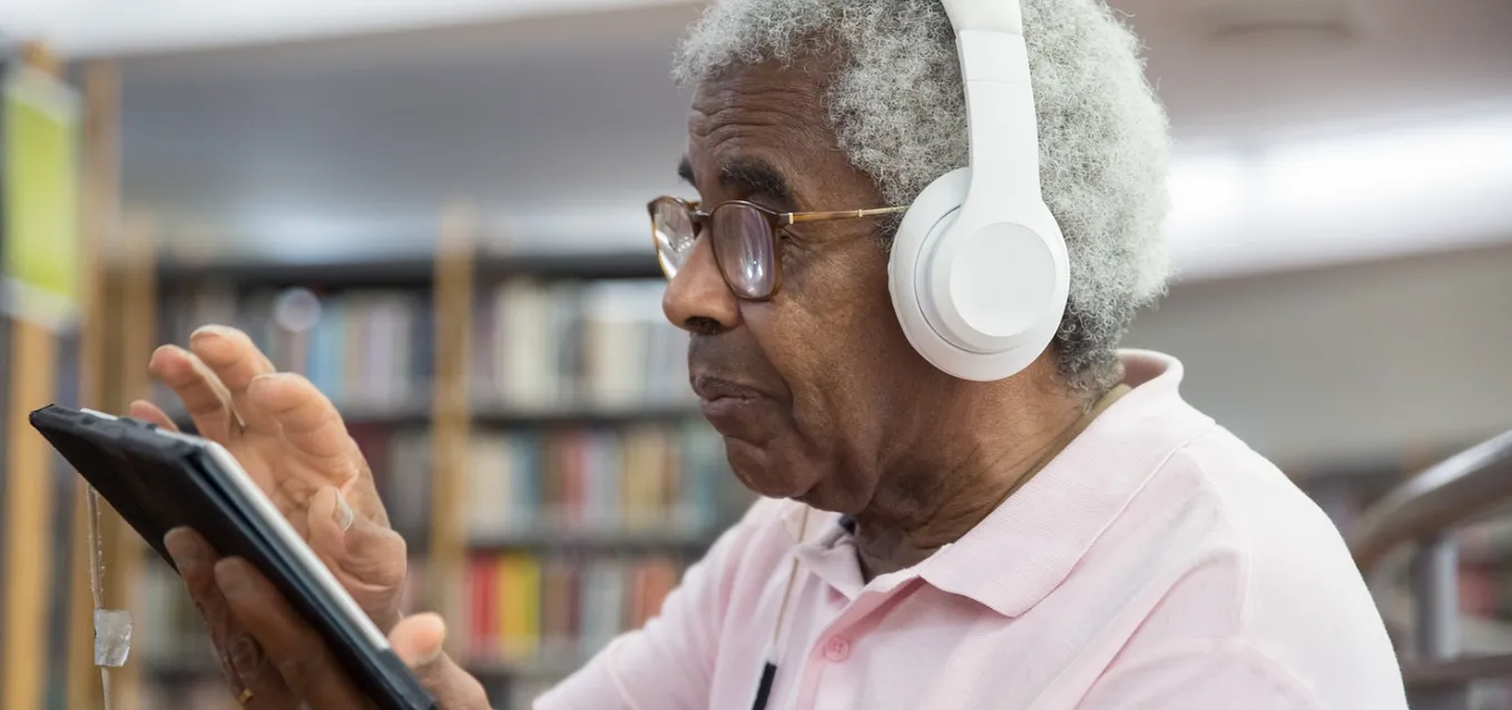 In the image, there is an elderly man who appears to be in his 70s or 80s. He is wearing headphones over his ears, and he is listening to something on his mobile or tablet device.