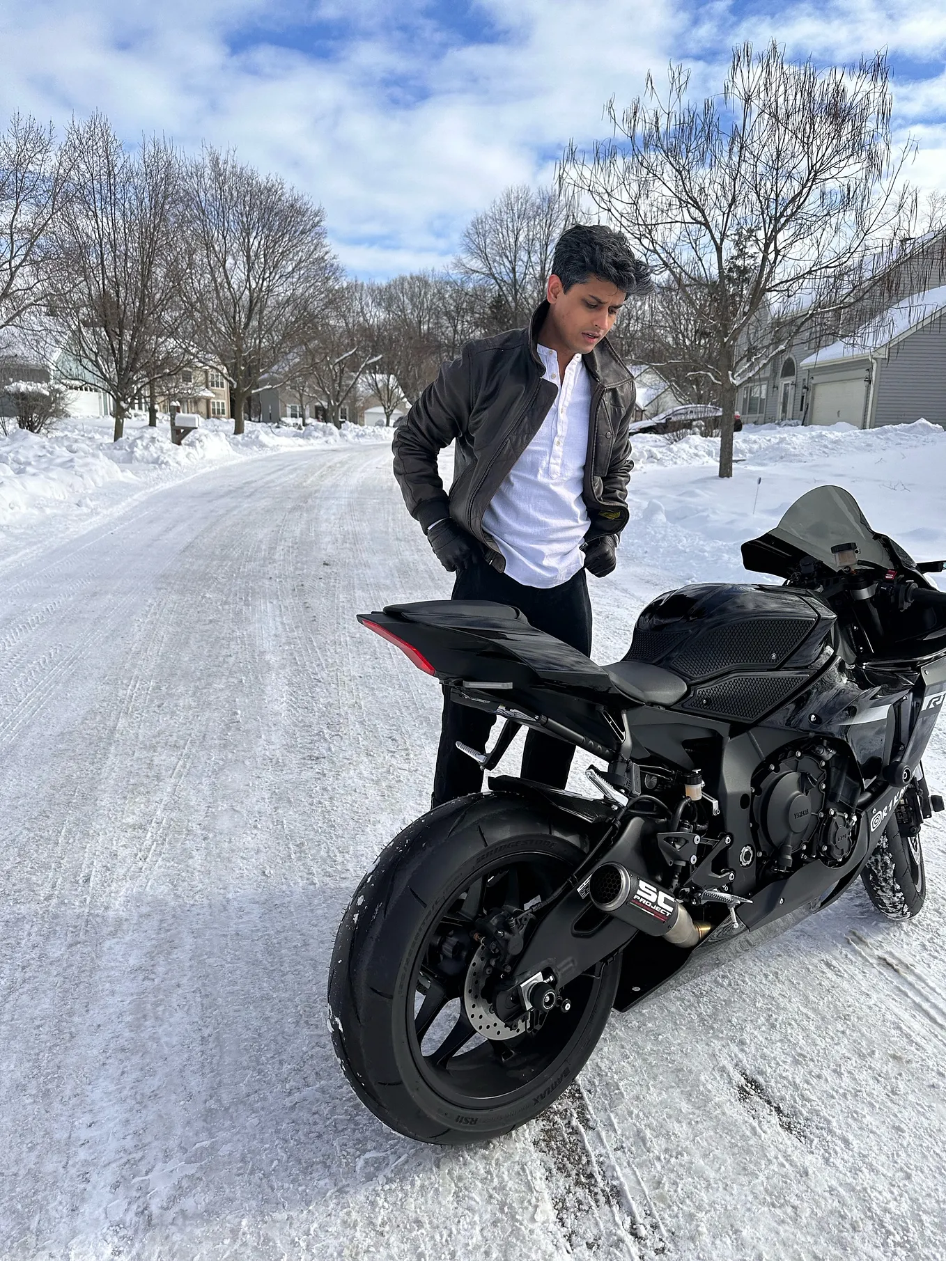 The photograph below captures a moment of quiet reflection: a man (myself) stands on a snowy street beside his sleek, black motorcycle. Dressed in a leather jacket and white shirt, he gazes downward, seemingly lost in thought, while the winter landscape stretches out behind him. The setting is serene, with bare trees lining the snow-covered road and the sky softly lit with clouds.