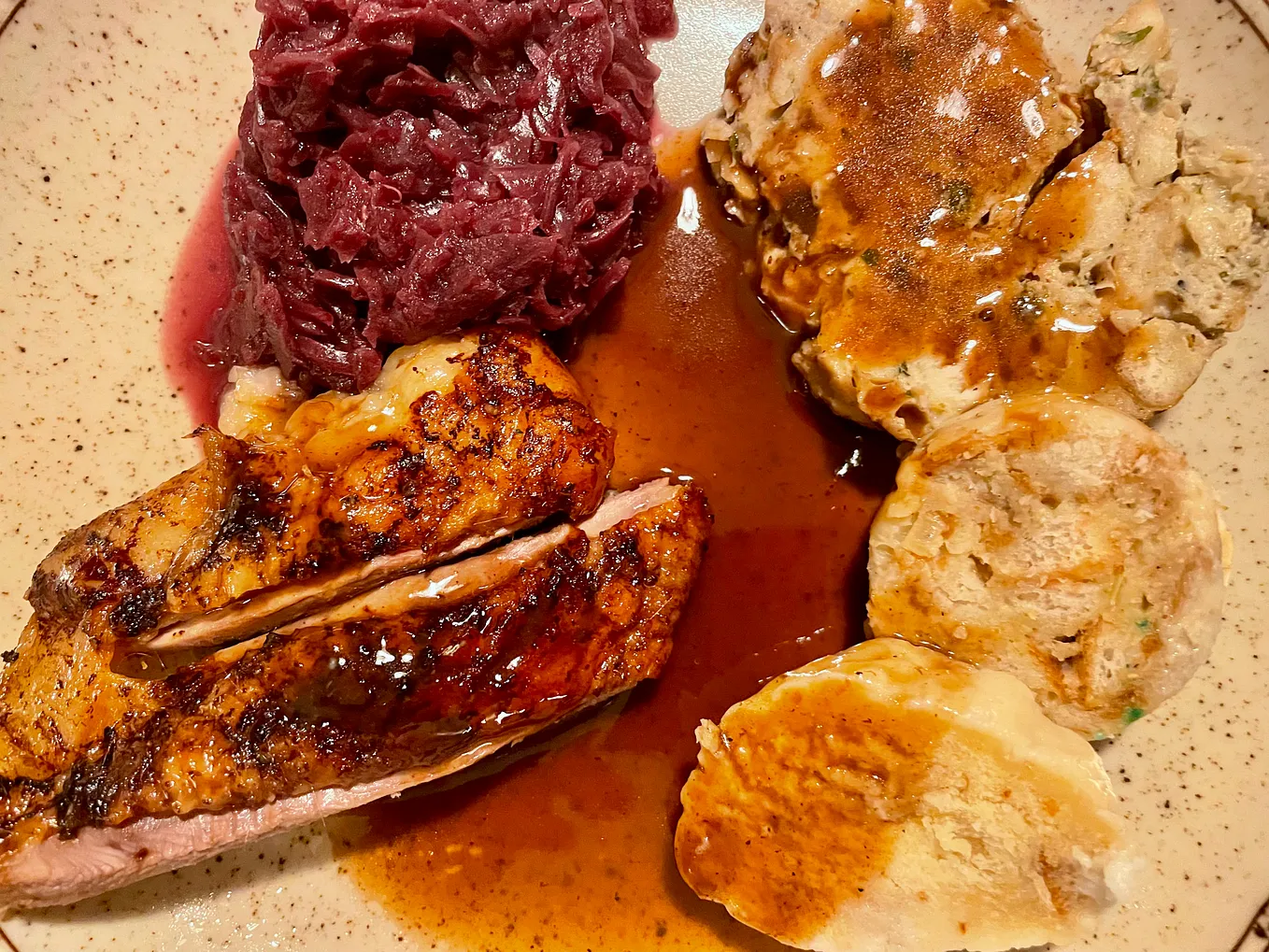 A close-up of a plate featuring sliced roasted goose with golden, crisp skin, covered in a glossy gravy. On the side are vibrant red cabbage and thick slices of Bohemian bread dumplings, all served on a speckled ceramic plate.