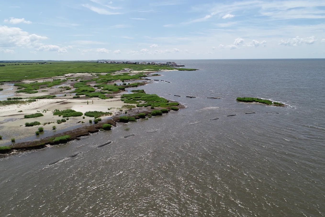 This aerial view shows the coastline with beach area and green marsh to the left. The ocean is to the right. In the water just off the sloping shoreline are breakwaters. There’s distance between each breakwater. A small peninsula no longer visibly connected to the coastline is visible. There is a row of residential structures along the water in the background.