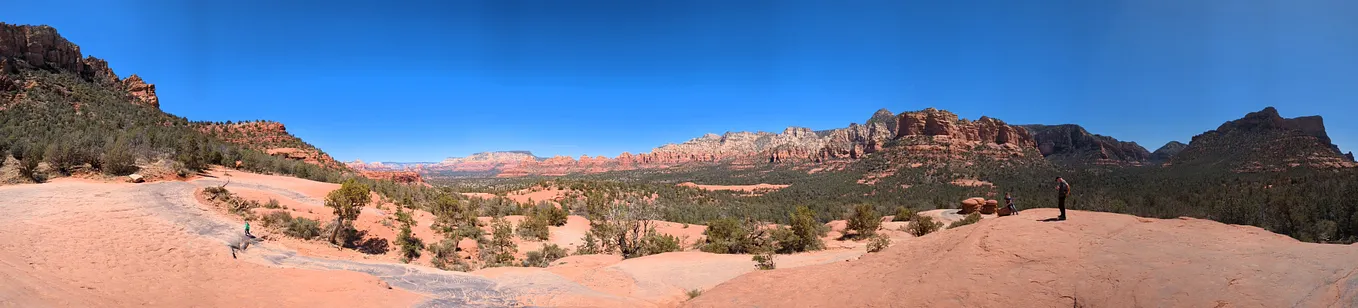 Distant mesas contrast against the bright blue sky and in the foreground a few tiny figures stand on large, round surfaces of red rock