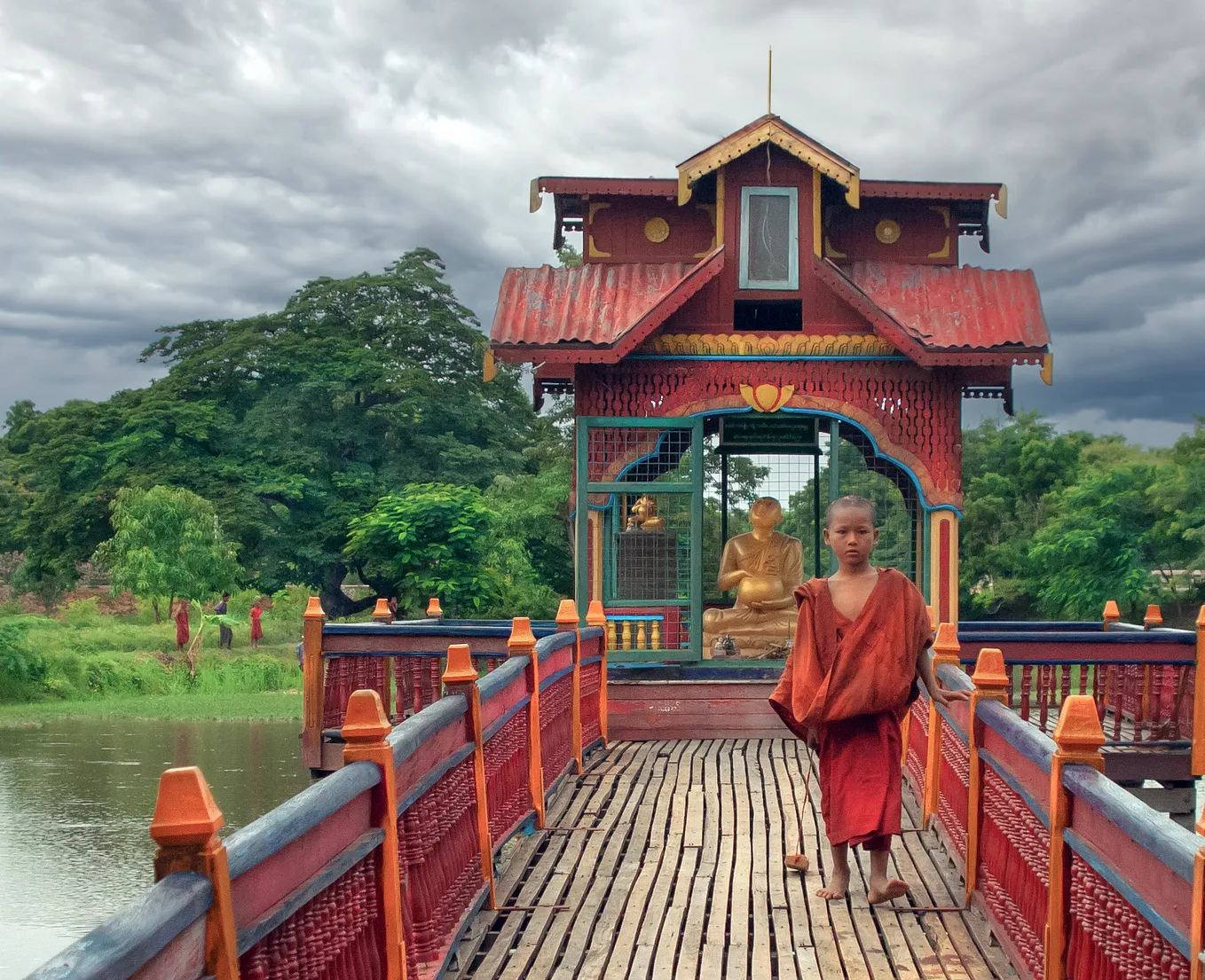 Young monk wearing an orange robe walks along a wooden bridge away from stupa where there is a golden Buddha sitting under the red titled roof.