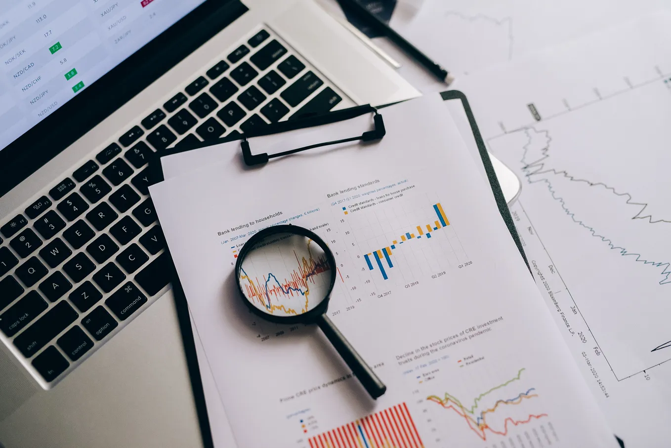 an open laptop with some papers showcasing data through graphs and visualization and a magnifying glass to take a deeper look into the graphs and data.