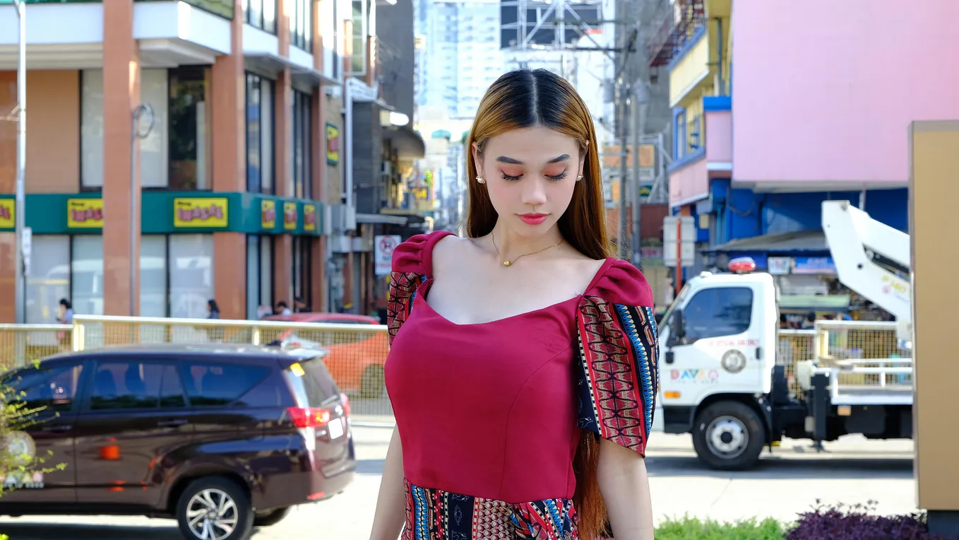 A woman wearing red Filipina Dress standing against a crowded San Pedro Street with shop houses and vehicles in the background.