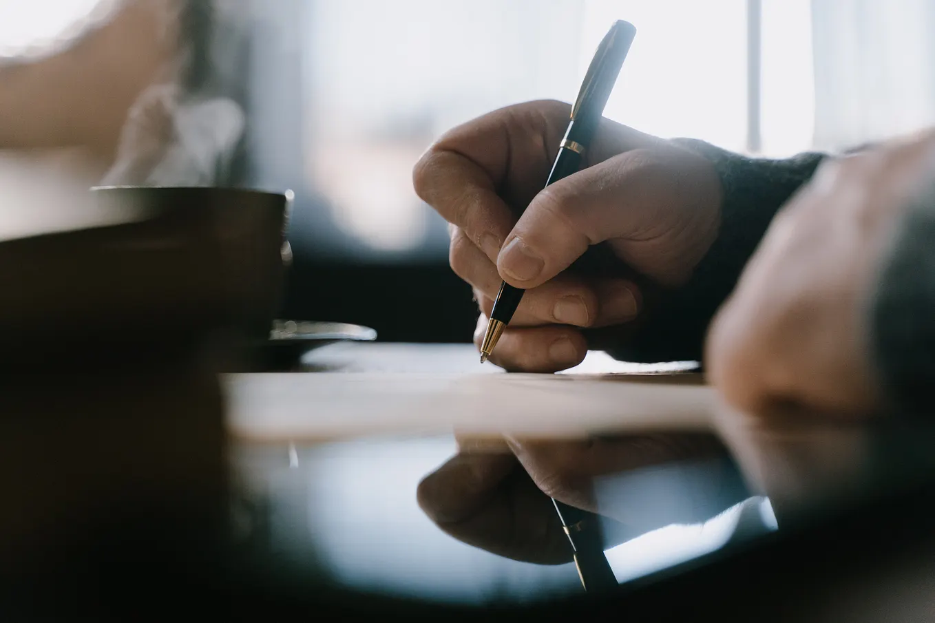 A side POV of hands holding a pen about to write on paper. Further back is a coffee cup with steam coming out of it.