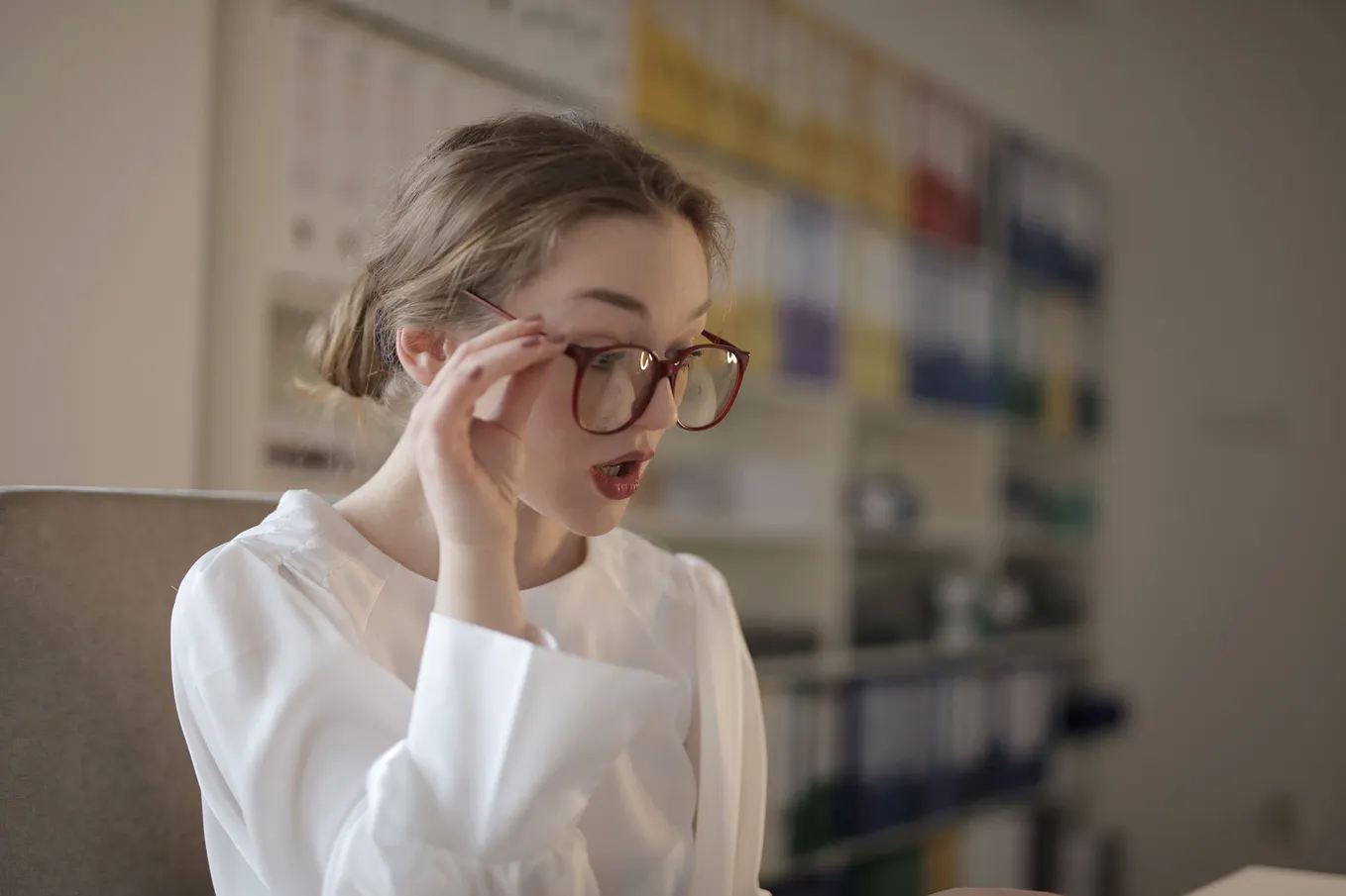 A woman in a white shirt holds a hand to her glasses with a shocked expression on her face