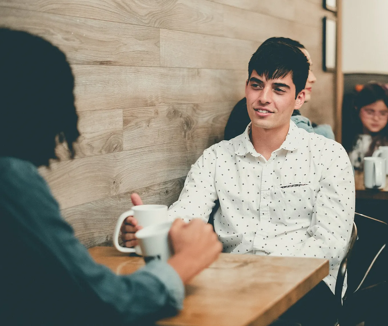 A man and a woman sitting at a table in a coffee shop, each holding acoffee cup.