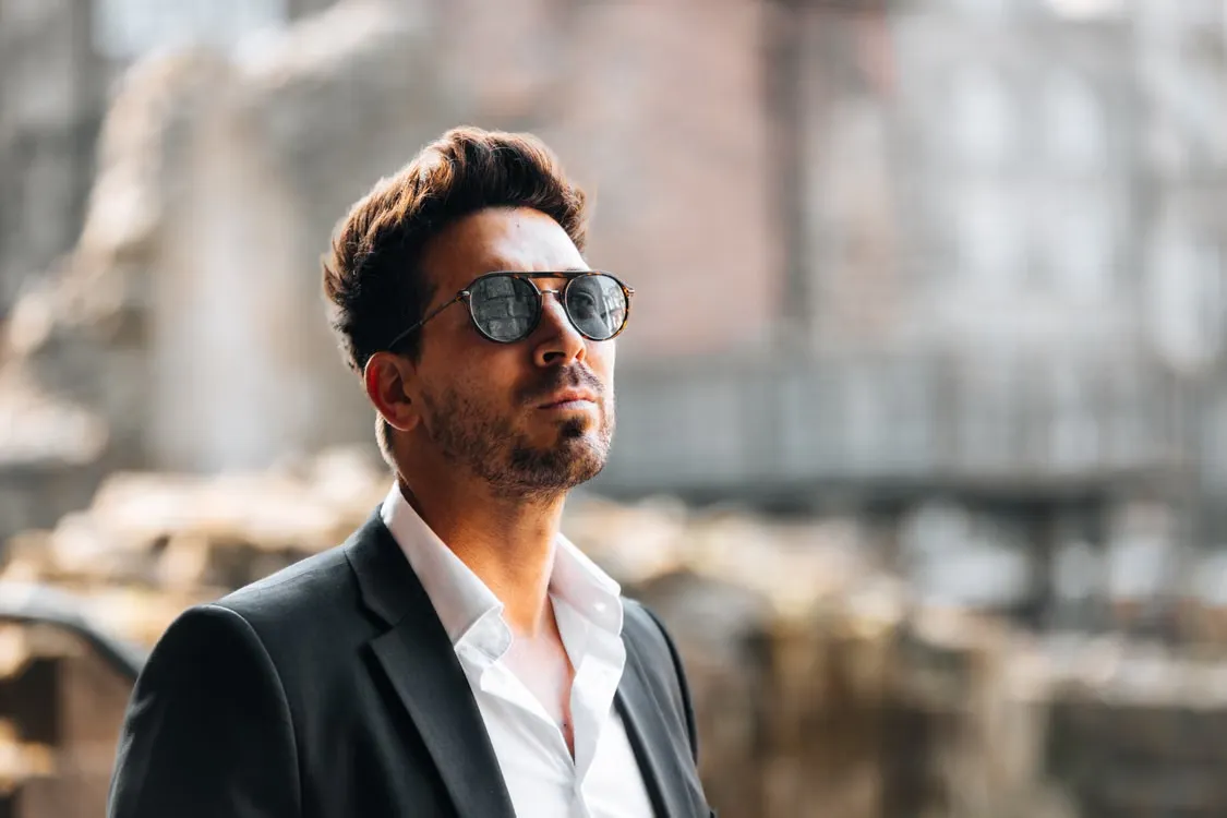A stylish man with sunglasses and a suit stands confidently outdoors in bright daylight