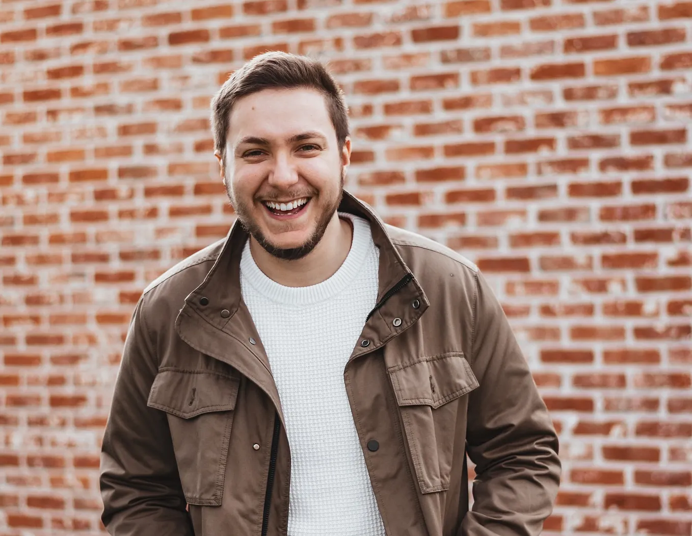 A young man smiling, in front of a brick wall.
