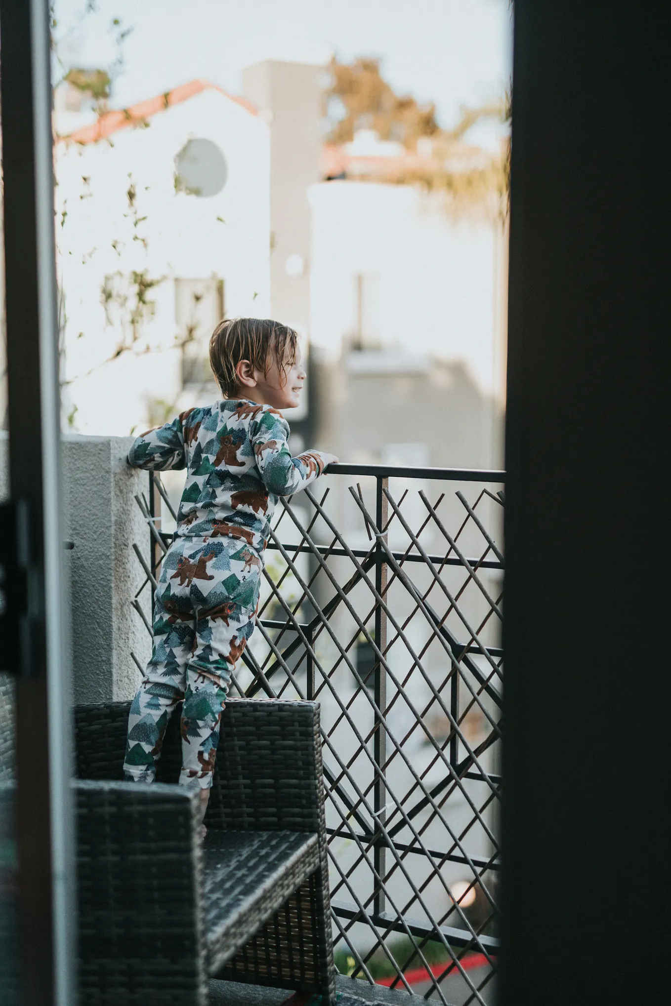 A little boy in splattered pyjamas standing on a box looking over a steel lattice railing into the day. He is about 5 or 6 years old with brown short hair. the view is from his back.
