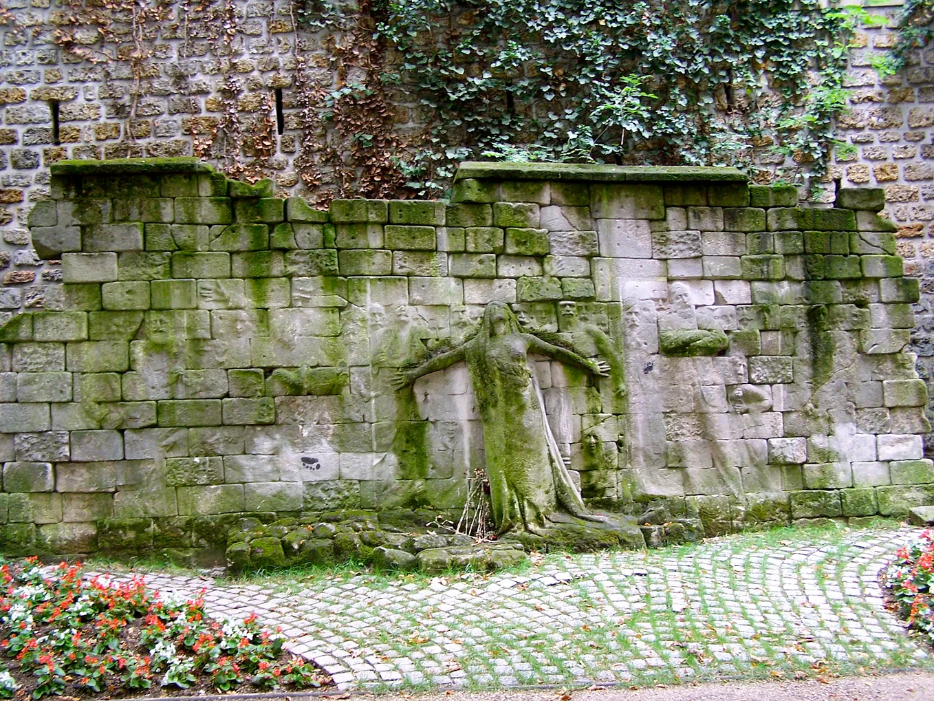 Carved in relief on an old stone wall is a plaintive female figure, arms outreached, eyes skyward, who can be clearly seen. Less visible are the much shallower reliefs, the carvings of people, mostly men with mustaches, who seem to hover around her.