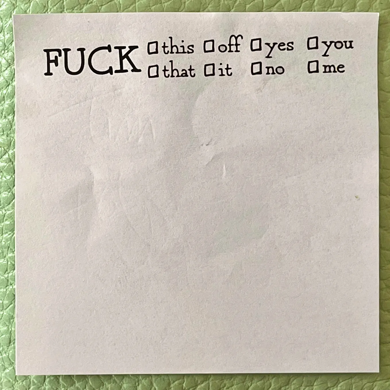 Color square image of a white post-it note with basic black font across the top of it stating in all-caps on the left side: “FUCK” with eight smaller lowercase font check boxes in two stacked rows to its right, the top row four boxes stating: “this, off, yes, you” and underneath that a row of box options stating: “that, it, no, me”. The post-it resting on a seafoam-green leather-skinned wallet.