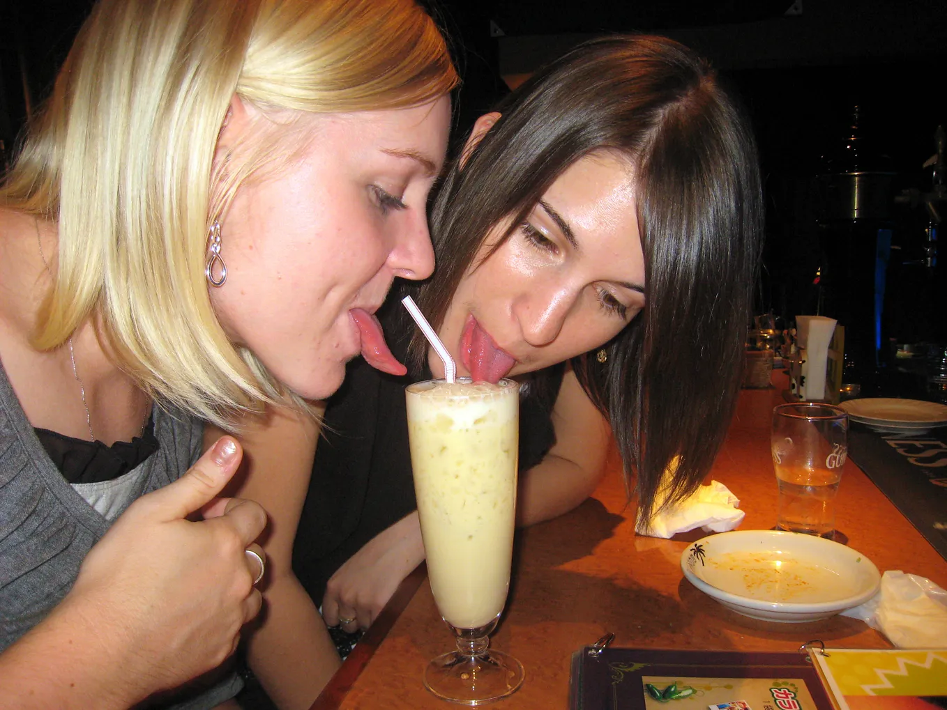 Two young women dip their tongues in a single glass of piña colada