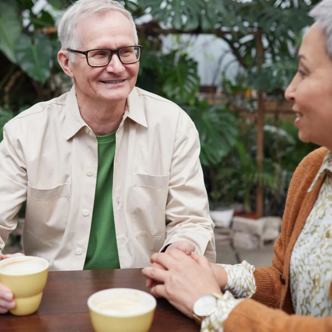 A gray haired gentleman has a cup of coffee and is holding hands with a woman amidst a setting of green trees and plants.