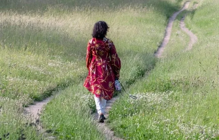 Young girl walking on a path through the grass, wearing a red patterned dress and white pants.