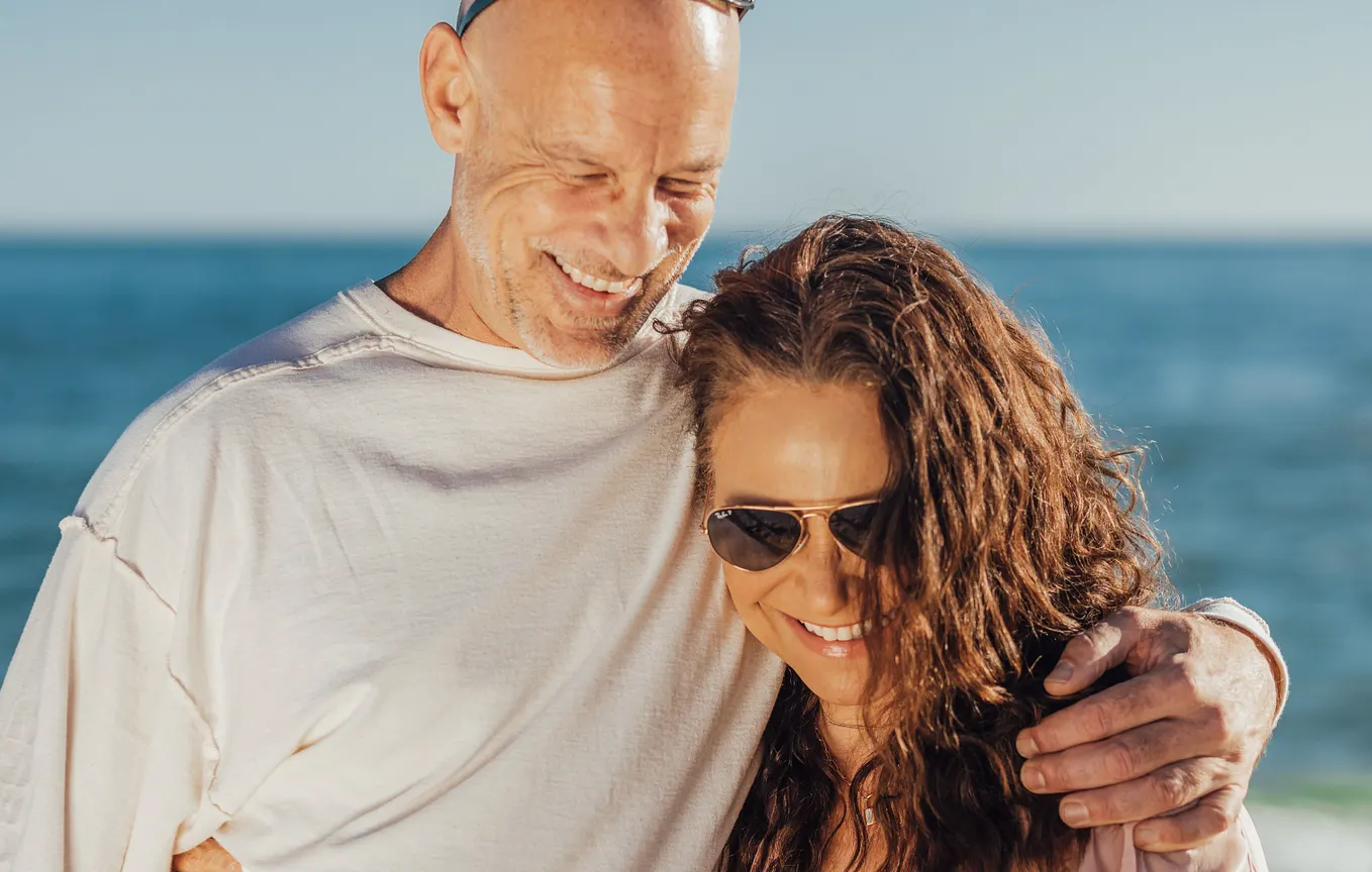 A smiling older man with his arm around a woman with long wavy brown hair and sunglasses on a sunny beach
