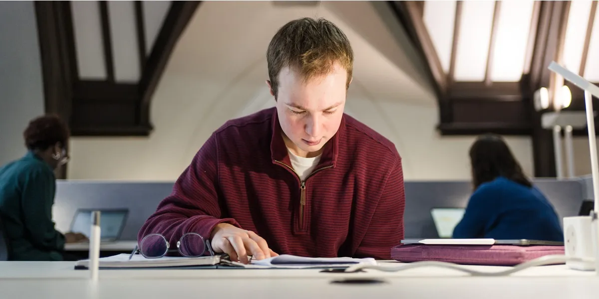 Jack sitting at a desk reading through his study materials. Two other students are sitting behind him working on their laptops at different desks.