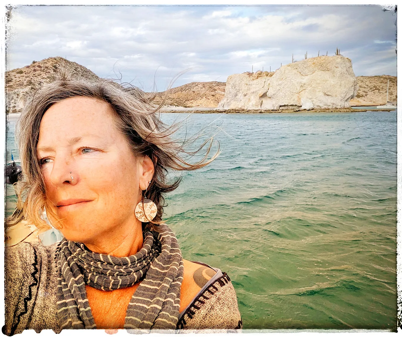 An older woman stares into the distance from a sailboat at anchor, wind-whipped waves and rocky cliffs in the distance.