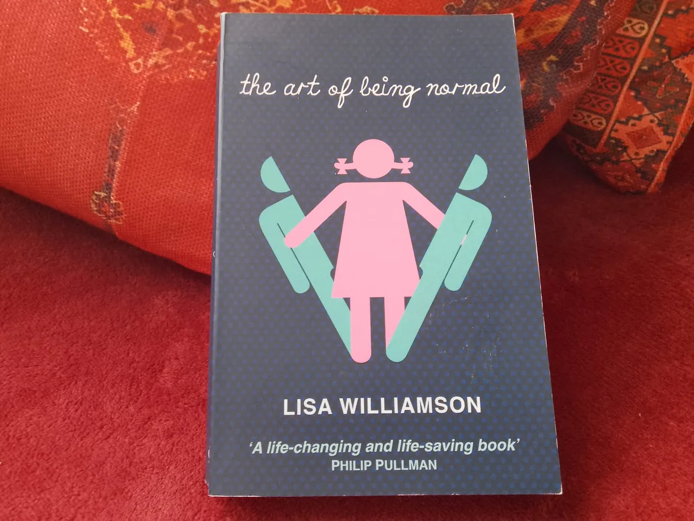 A photograph of the author’s copy of Lisa Williamson’s novel “The Art of Being Normal”. It’s the David Fickling edition, published in 2015, and the artwork shows an image based on the generic male/female symbols used to indicate gender-specific bathrooms, with a female figure emerging from a male figure breaking in two.