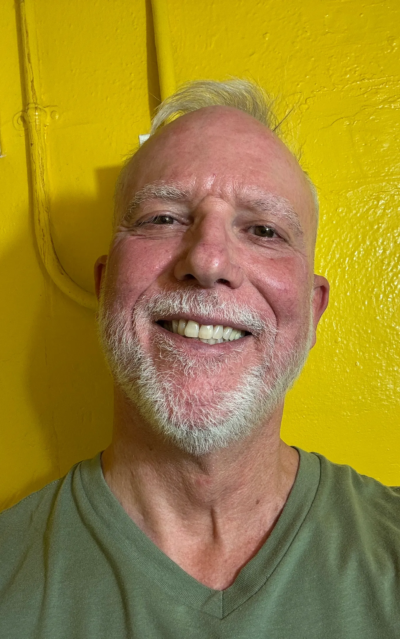 Self-Portraint of the author, a smiling silver coloured bearded man, stood against a striking yellow background.