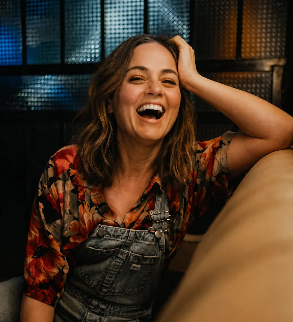 A Gen X woman in casual overalls laughing while relaxing on a couch.