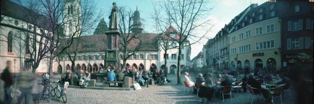 A paronamic photo of a square in Freiburg (Germany) with a fountain, tables and many people sourounded by old architecture.