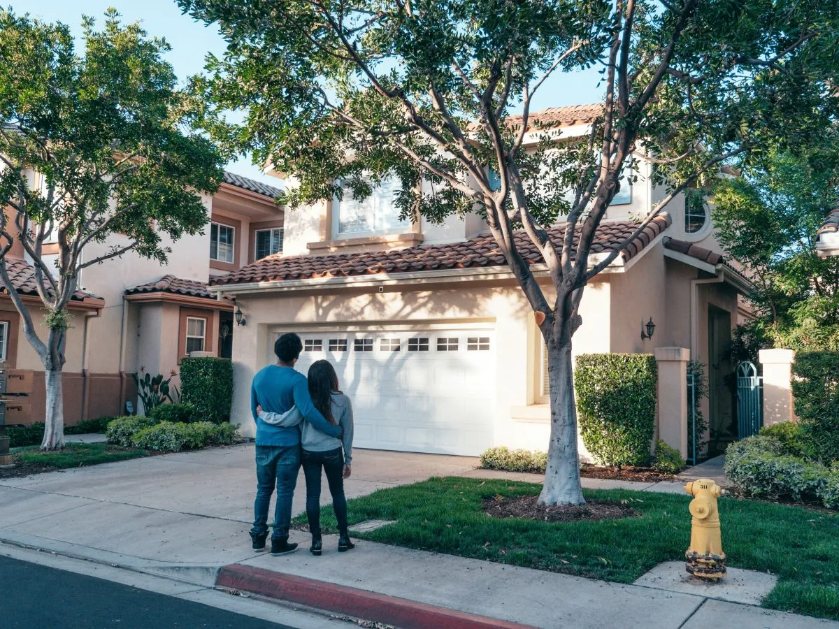 A young couple standing, arms around each other, looking at a suburban house.