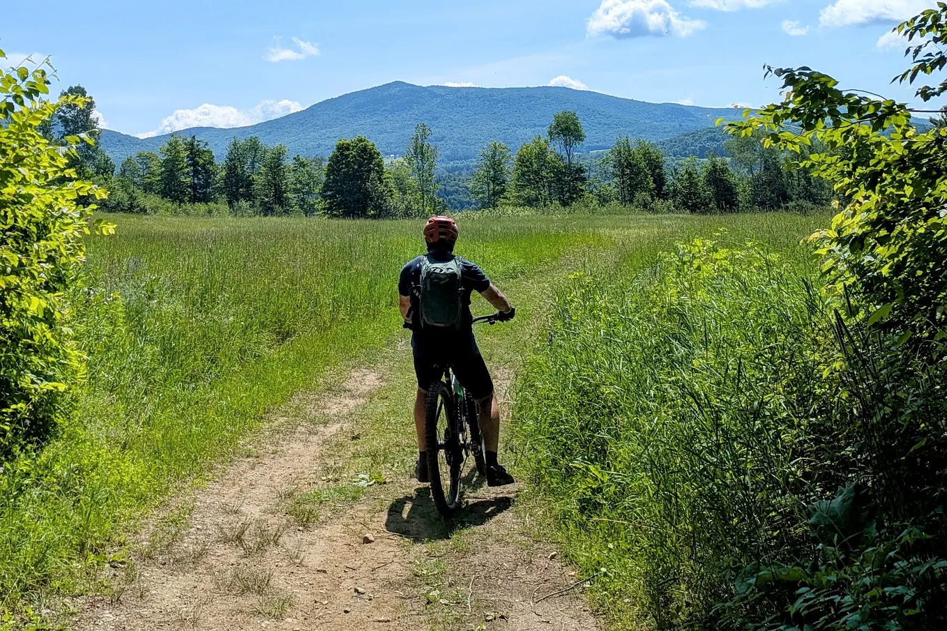 The author on a mountain bike, stopped on a trail through a green field, looking out at Burke Mountain on a sunny day.