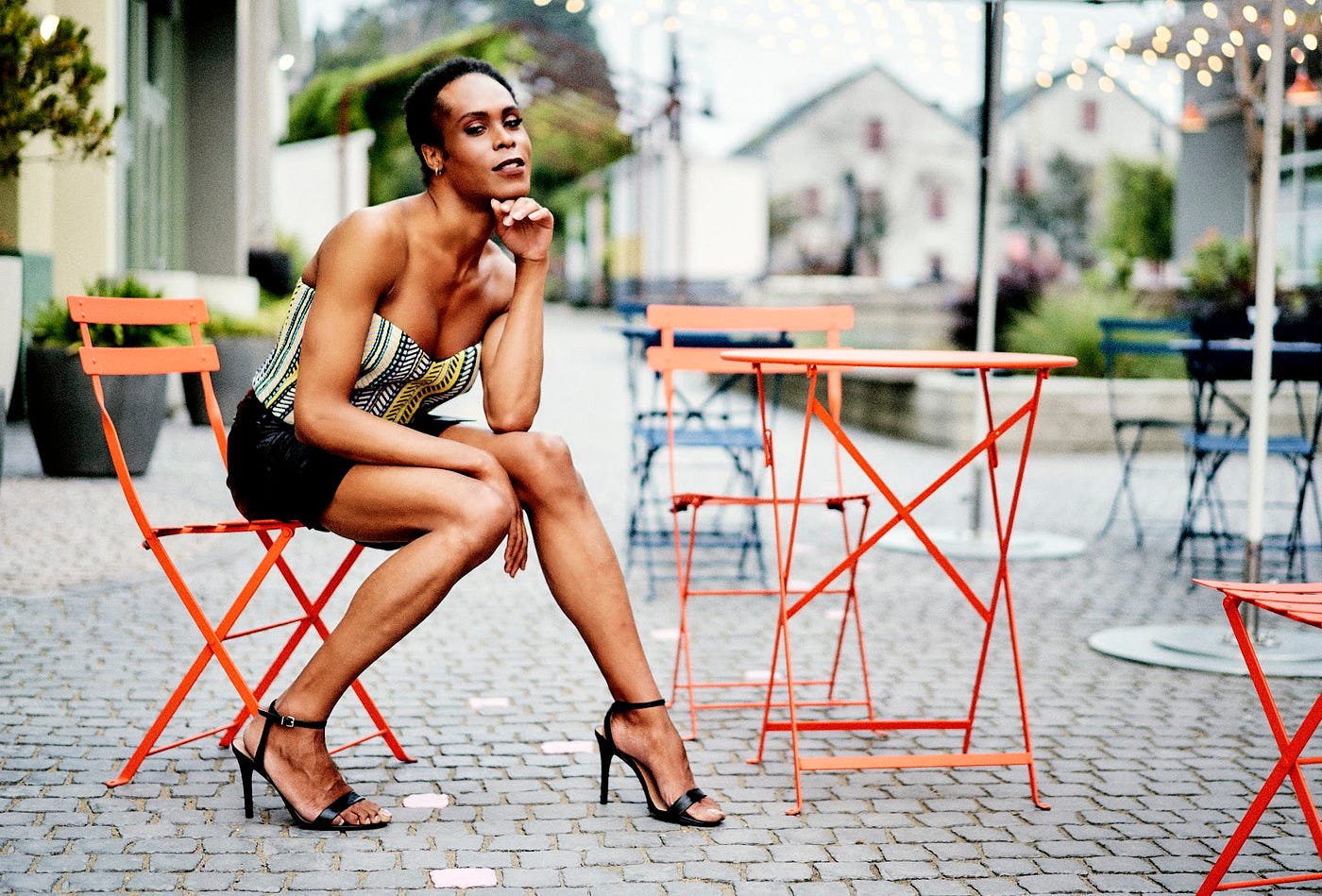 A Black woman with very short hair, a sleeveless shirt, a short skirt, and strappy heels sitting at an outdoor dining space.