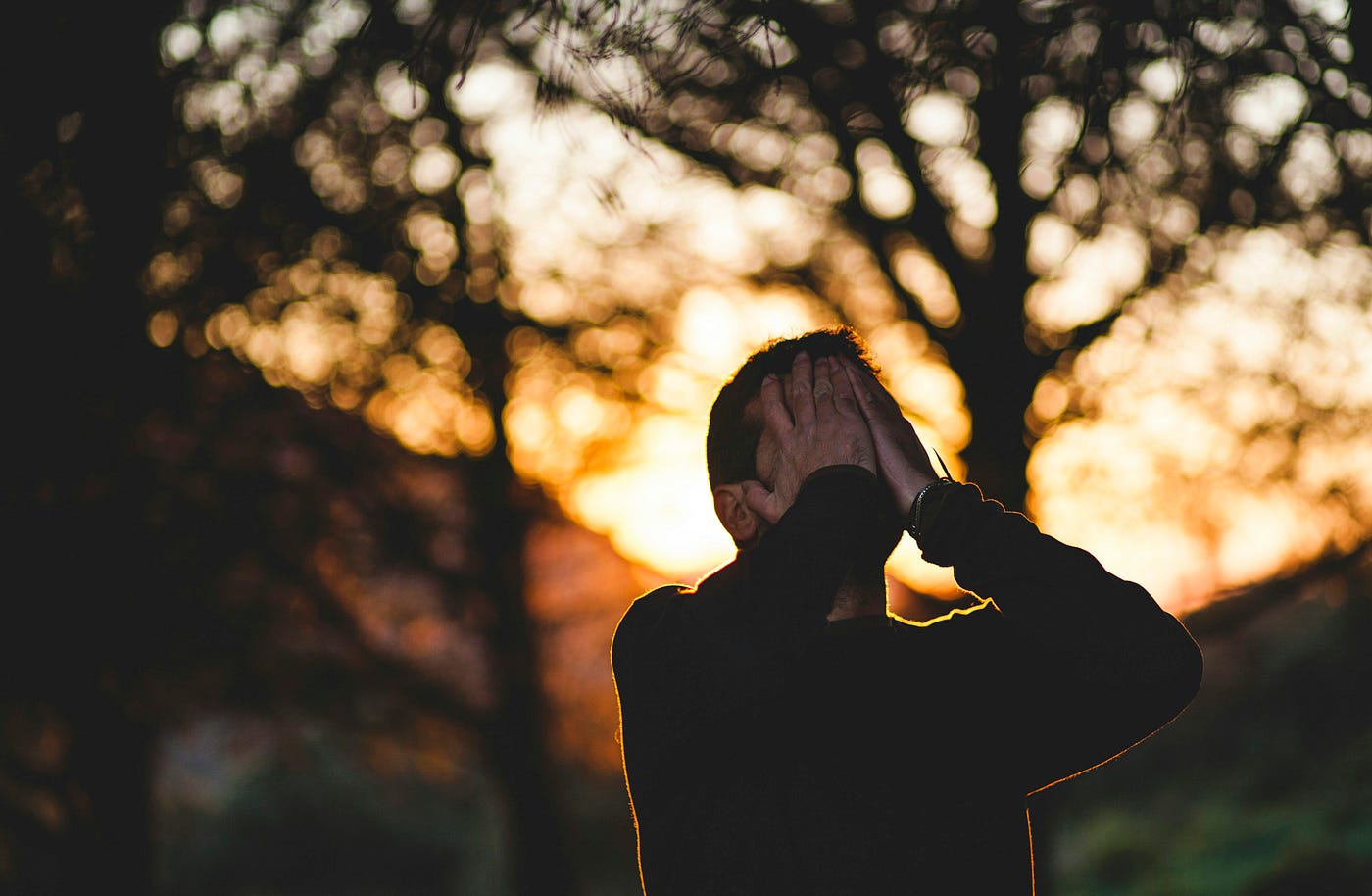 A young man puts his hands on his face in a gesture of feeling stressed. There are blurry trees in the background.