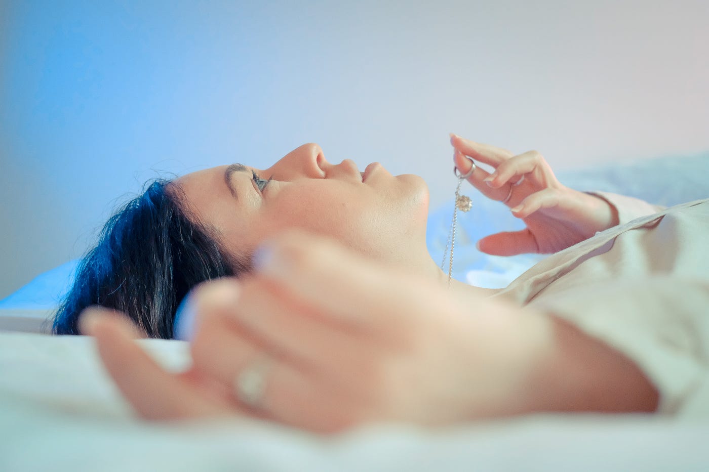 A young woman lies with her head to the left, her arms raised to ear level. She appears to be nearly floating on a bed as she gases upward. Lack of sleep or poor sleep quality can lead to various health issues, including increased risk of chronic diseases, impaired cognitive function, weakened immune system, and compromised emotional well-being.