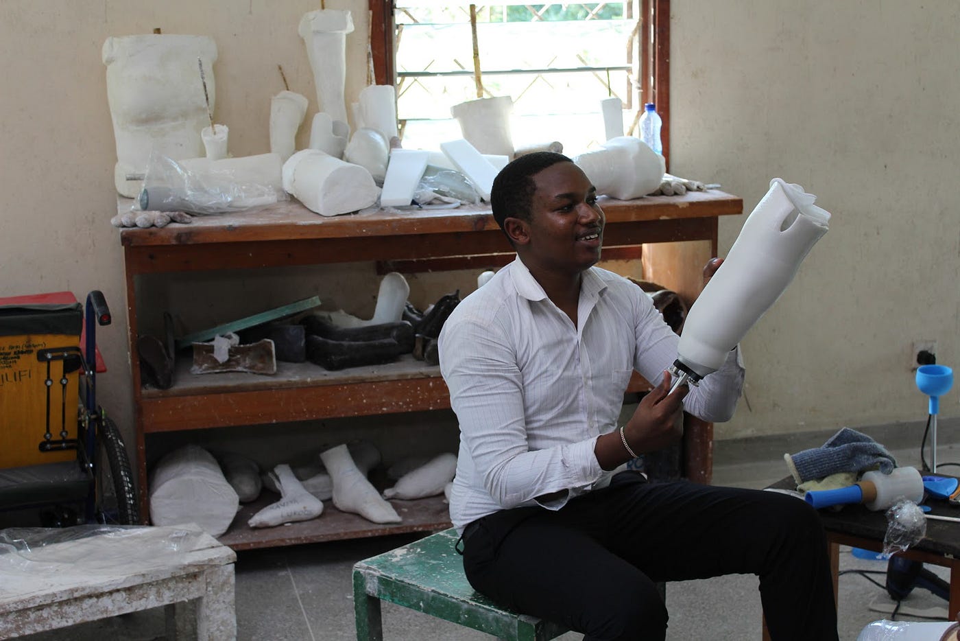 A young black entrepreneur is smiling as he holds up a white prosthetic limb to the light. Various other prototypes behind.