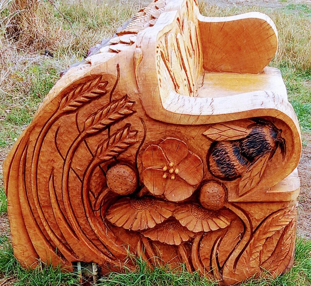 Image of the side of a bench carved from a tree trunk, depicting a bee, some flowers and some wheat sheafs, to illustrate post