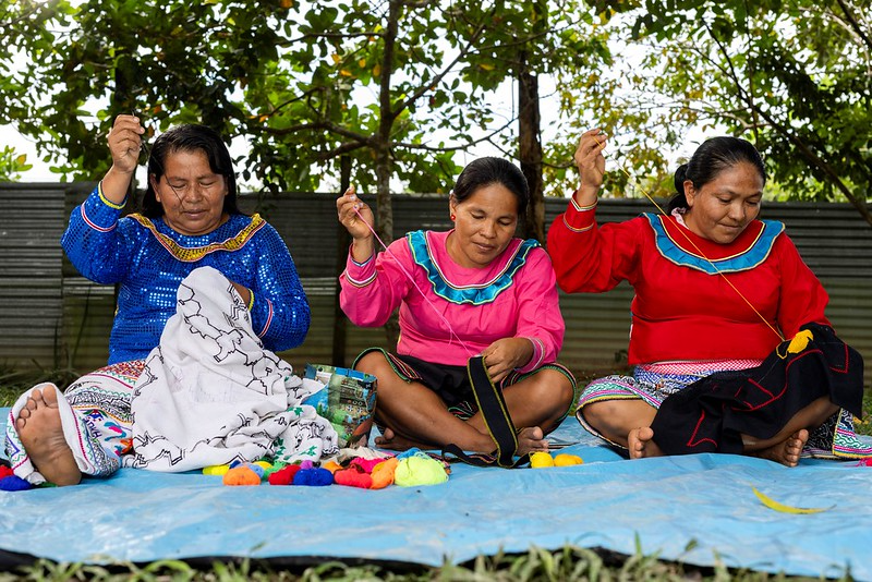 Three Shipibo-Conibo women artists sit on the ground while sewing traditional patterns into textiles.
