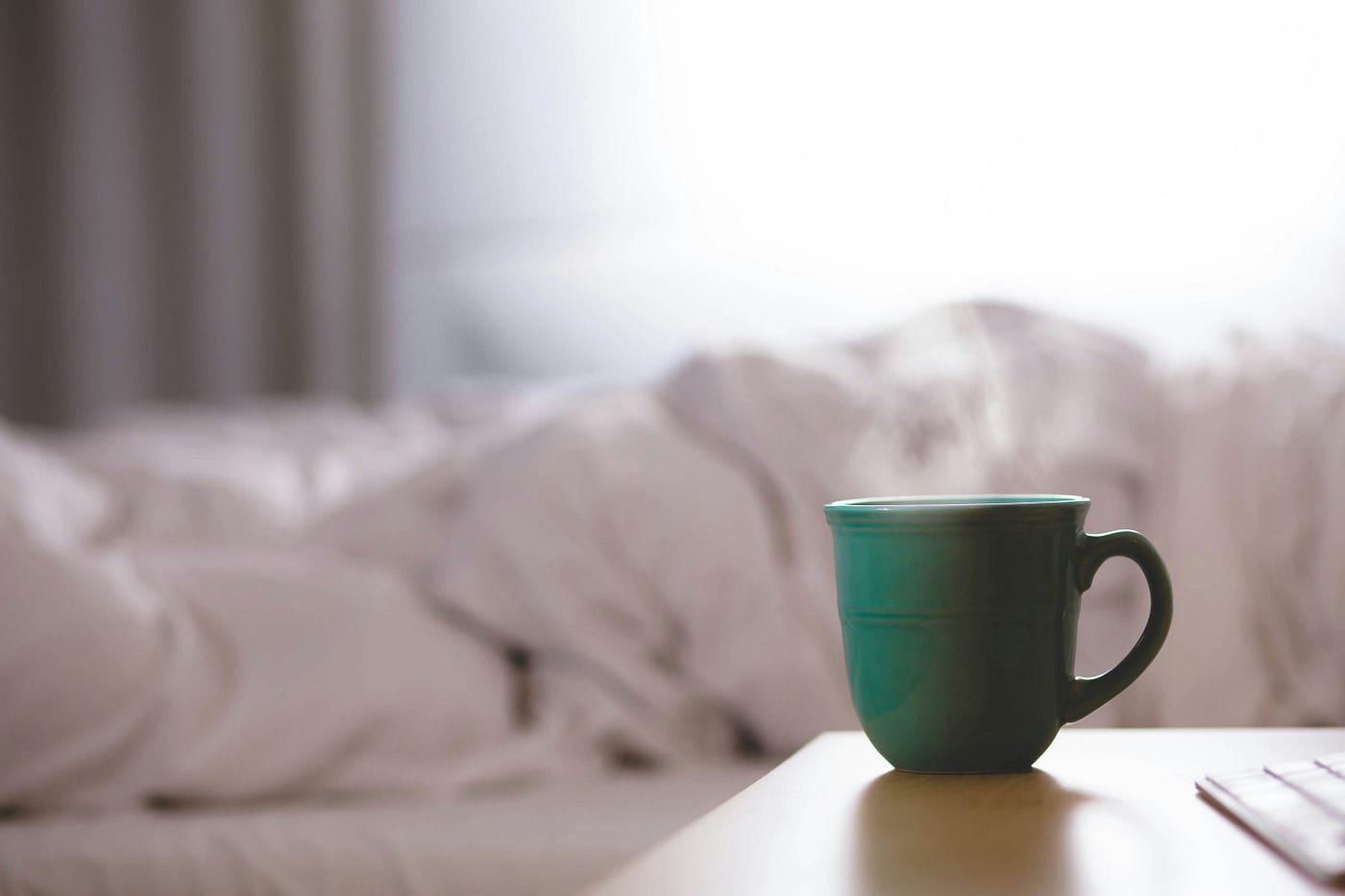 A steaming green coffee cup sits on a desk, with a a bed and a white comforter in the background.