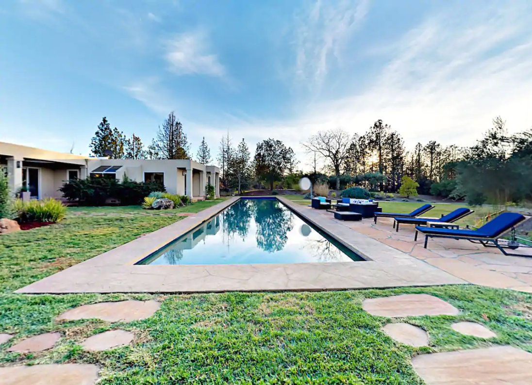 A large rectangular pool flanked by a row of blue lounge chairs on one side and squarish single-story house on the other.