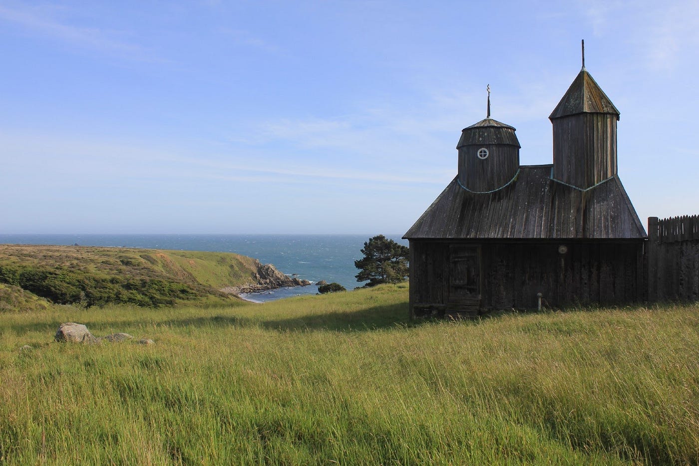 The wooden Orthodox church in the first picture, seen from the other side of its fence in a meadow, ocean in the background.