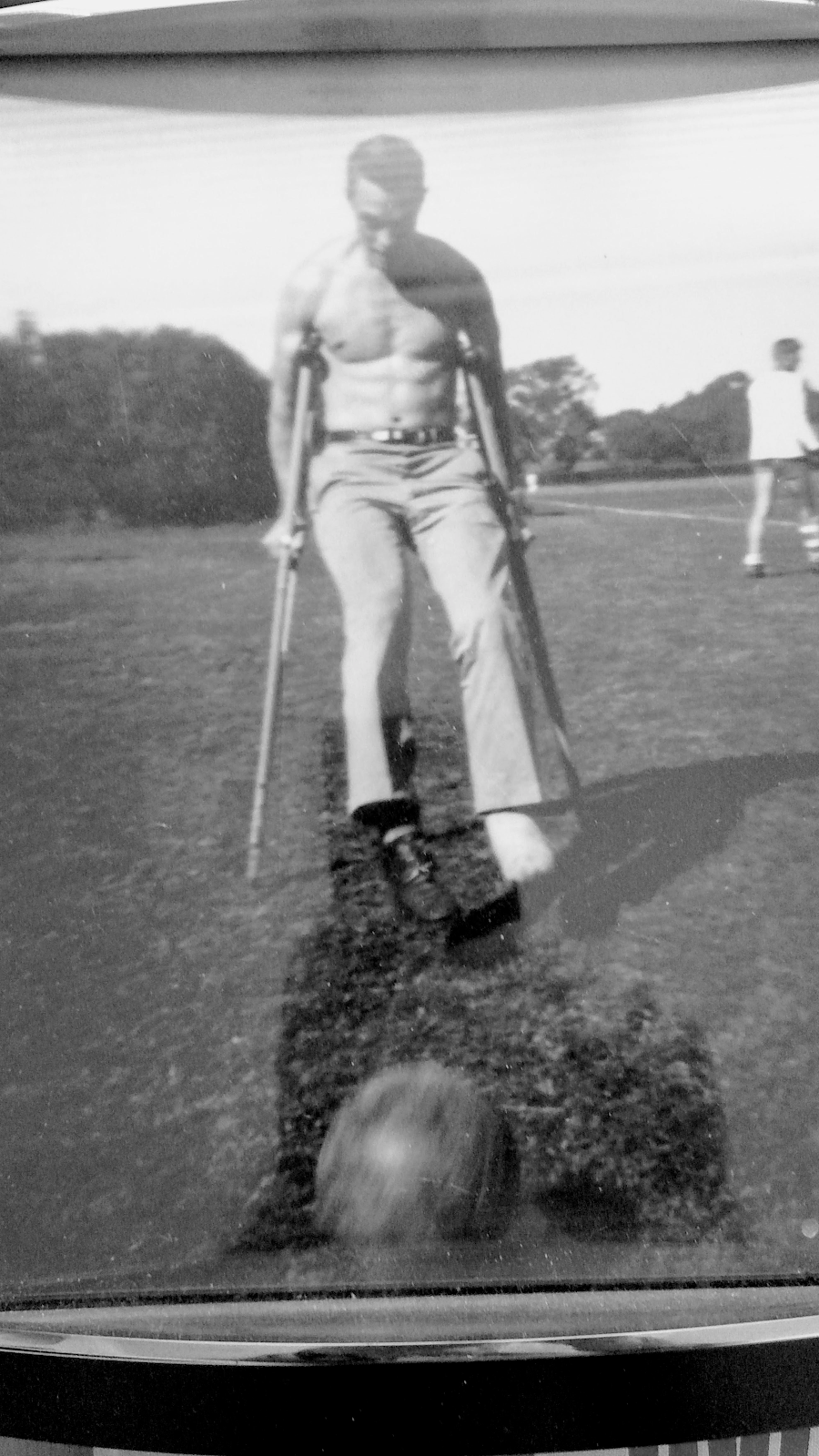Black-and-white photo of a shirtless man using crutches on a field, one foot bandaged, to kick a soccer ball.