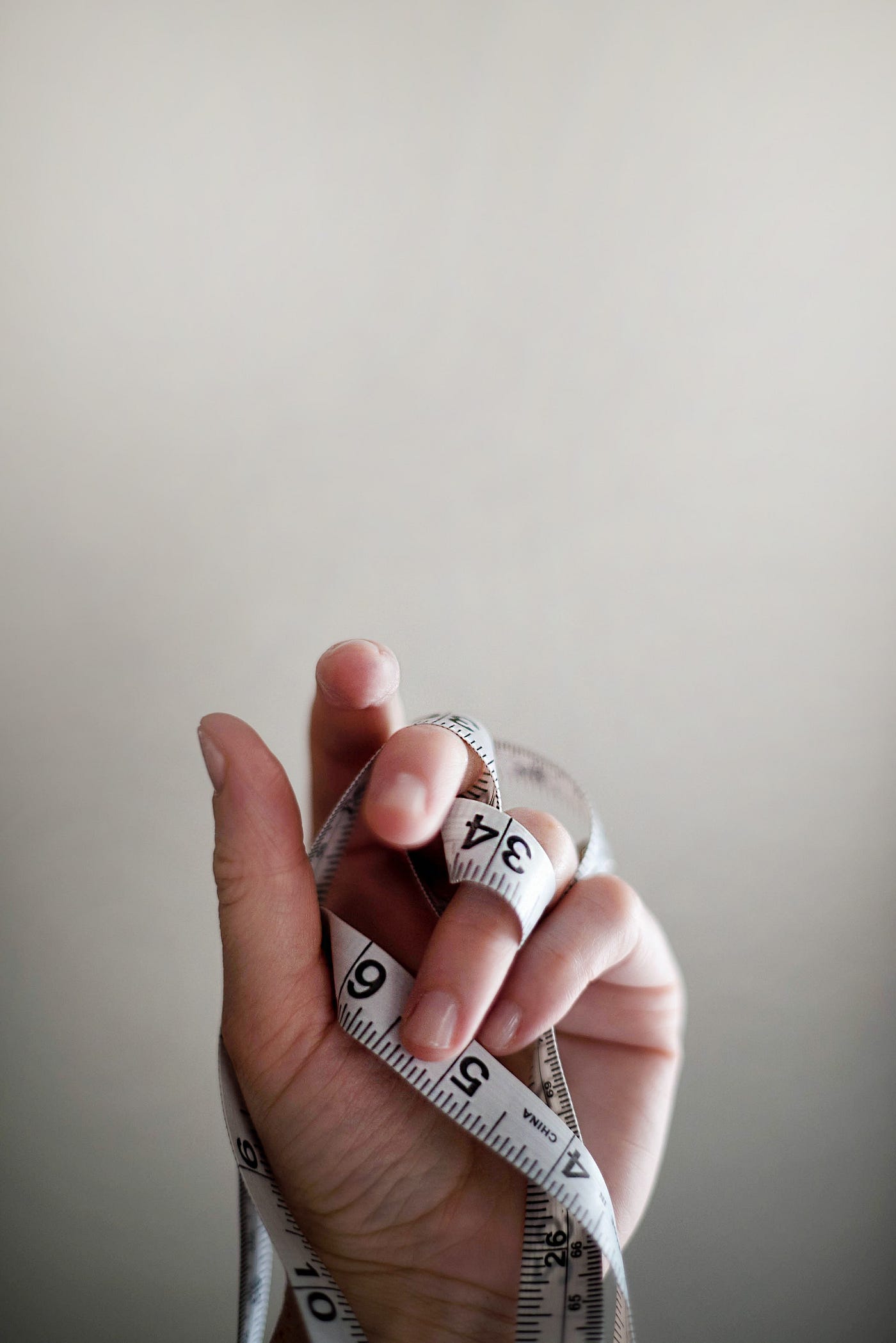 A hand reaches up, holding a paper tape measure that is intertwined with the person’s fingers on the left hand.