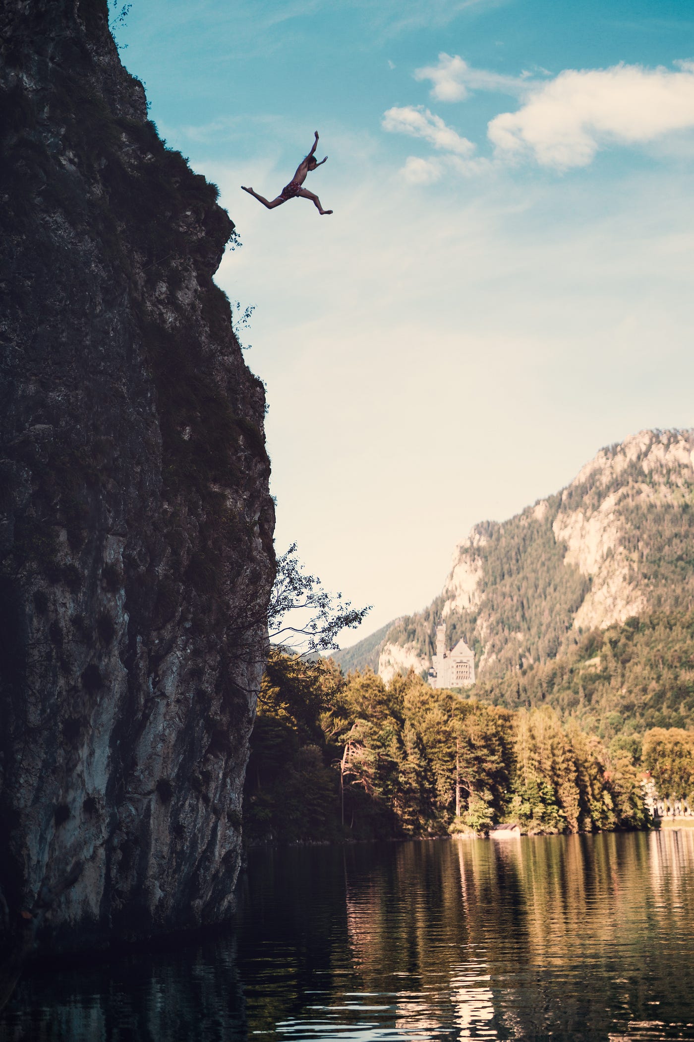 A person jumping off of a tall cliff on camera left into a lake with mountains surrounding.