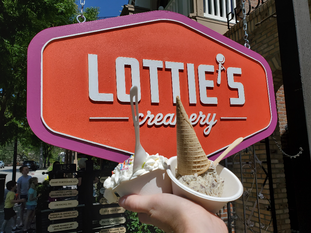 A hand holding two single-cup cones in small bowls in front of the Lottie’s Creamery exterior sign.