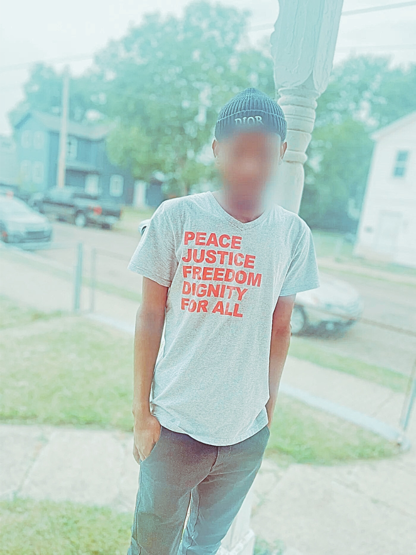 A young man with a blurred face wears a t-shirt that says “Peace, Justice, Freedom, Dignity for All.”