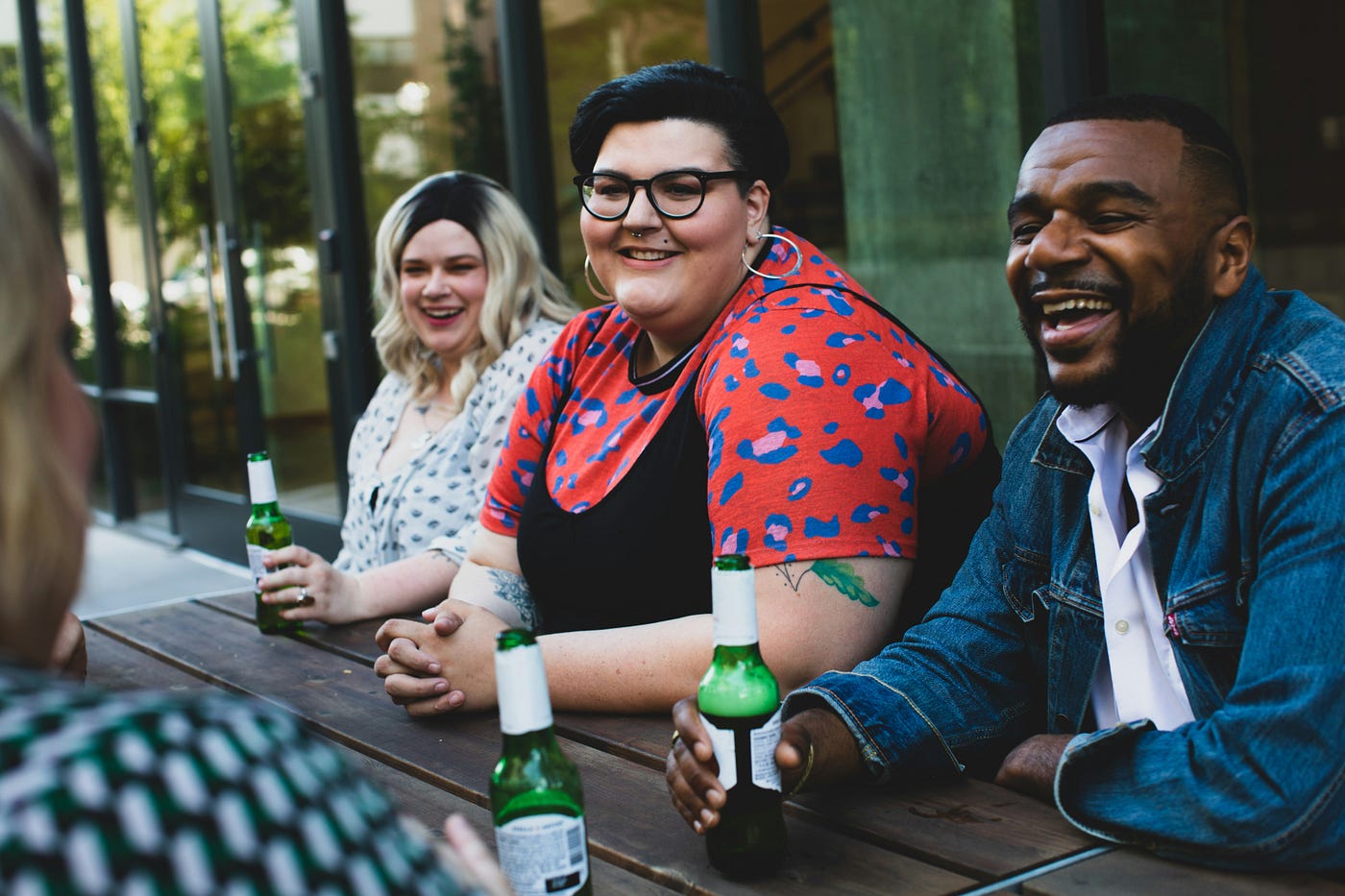 Three young individuals smile as they sit at a picnic table, drinking beers.