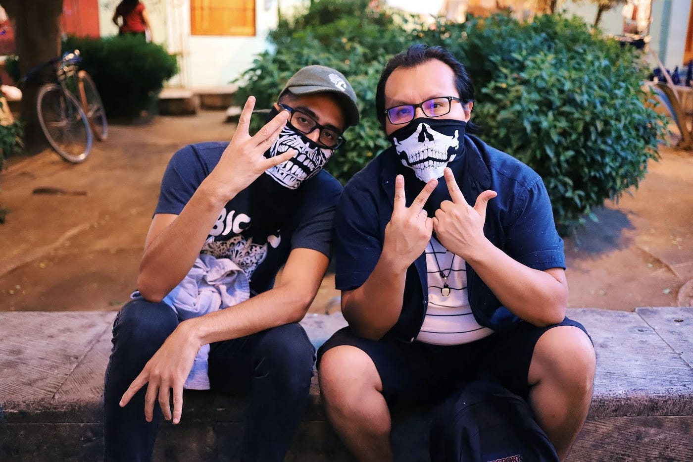 Two people wearing skull gaiters on their faces, holding fingers in W shapes, sitting on a stone bench in front of a house at night.