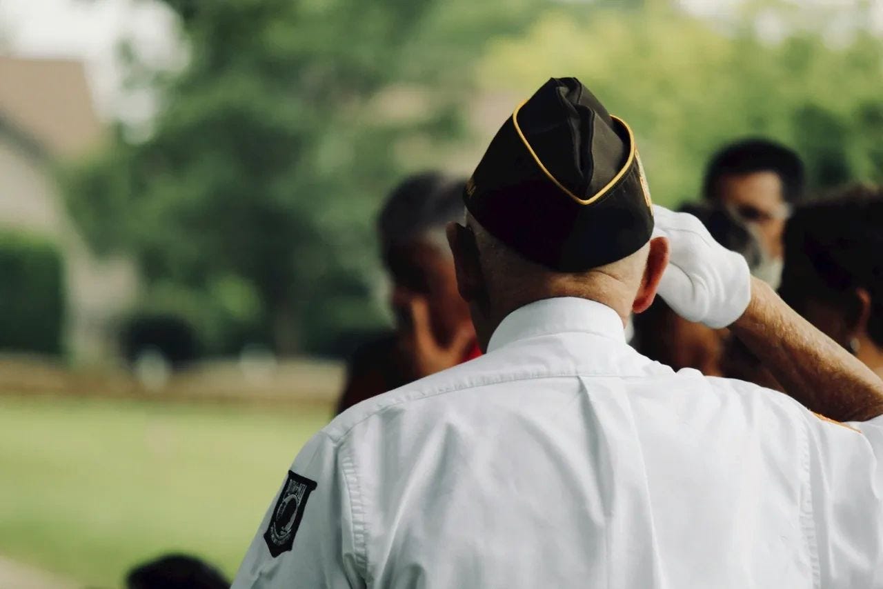 Mickey Markoff Air Sea Exec 2024 — photo of man in military uniform with POW patch on arm saluting crowd. Posted on 2024 Mickey Markoff article on net worth of paying tribute to our nation’s heroes.