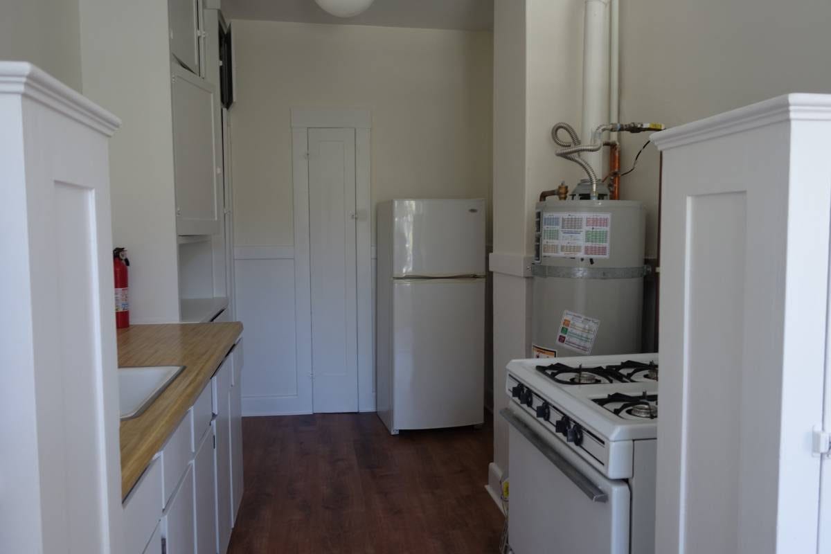 Cabinets, a sink, an oven, and a water heater in the foreground, a fridge lurking in the background next to a skinny door.