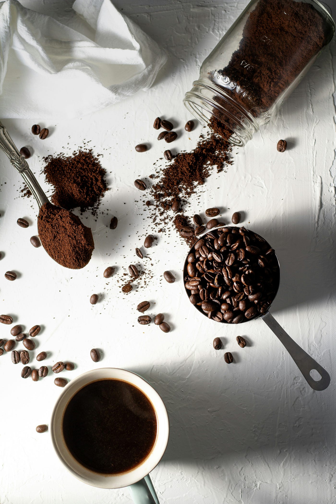 A coffee cup, spoon filled with coffee beans, a smaller spoon with ground coffee, and a glass Mason jar on its side (spilling ground coffee).