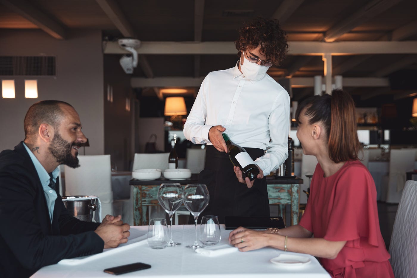 A waiter wearing a face mask shows a bottle of red wine to a maskless couple in a restaurant.