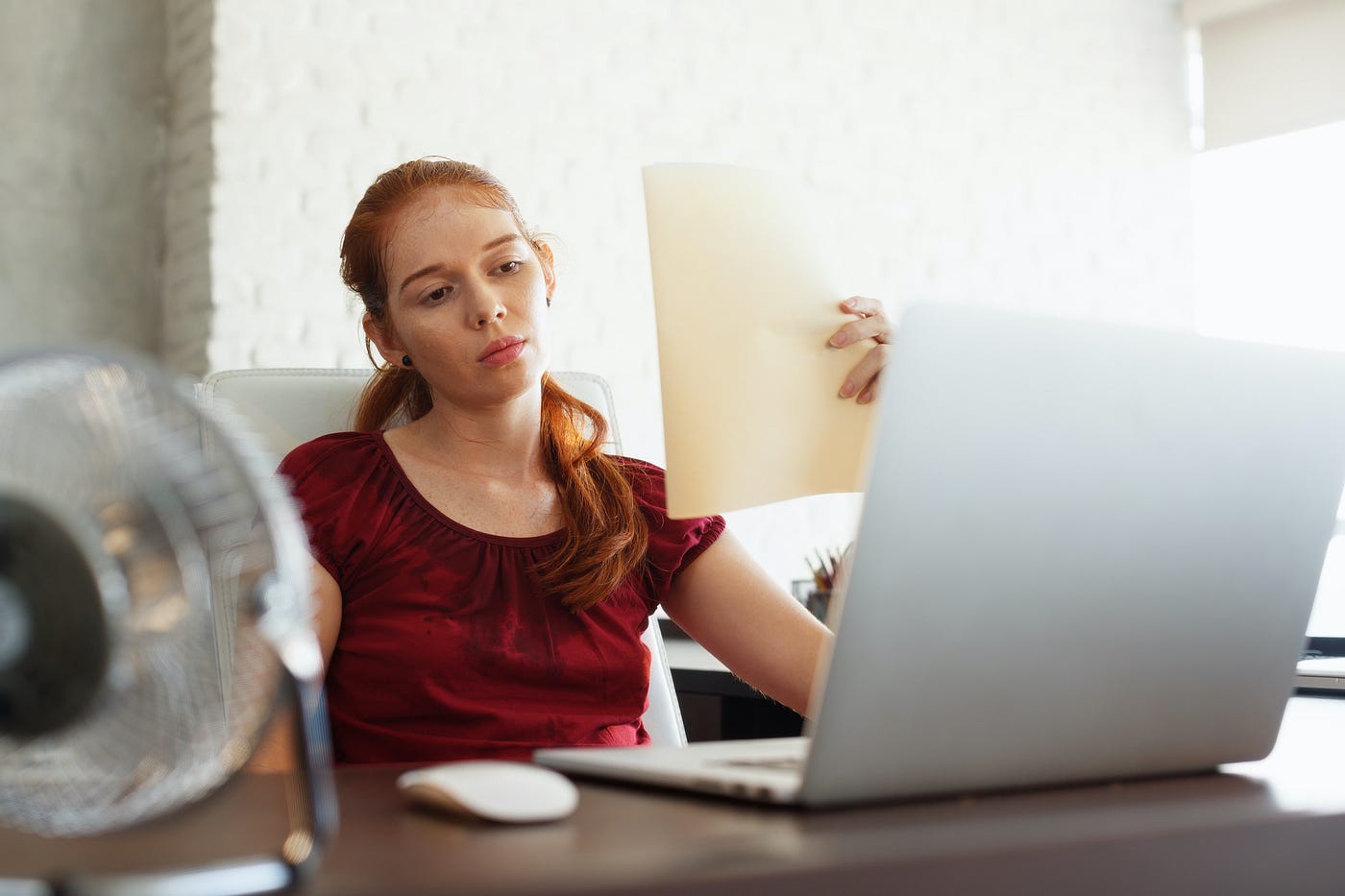 A person trying to work from home in a heatwave. A fan is pointed at their face and they’re fanning themself with paper.