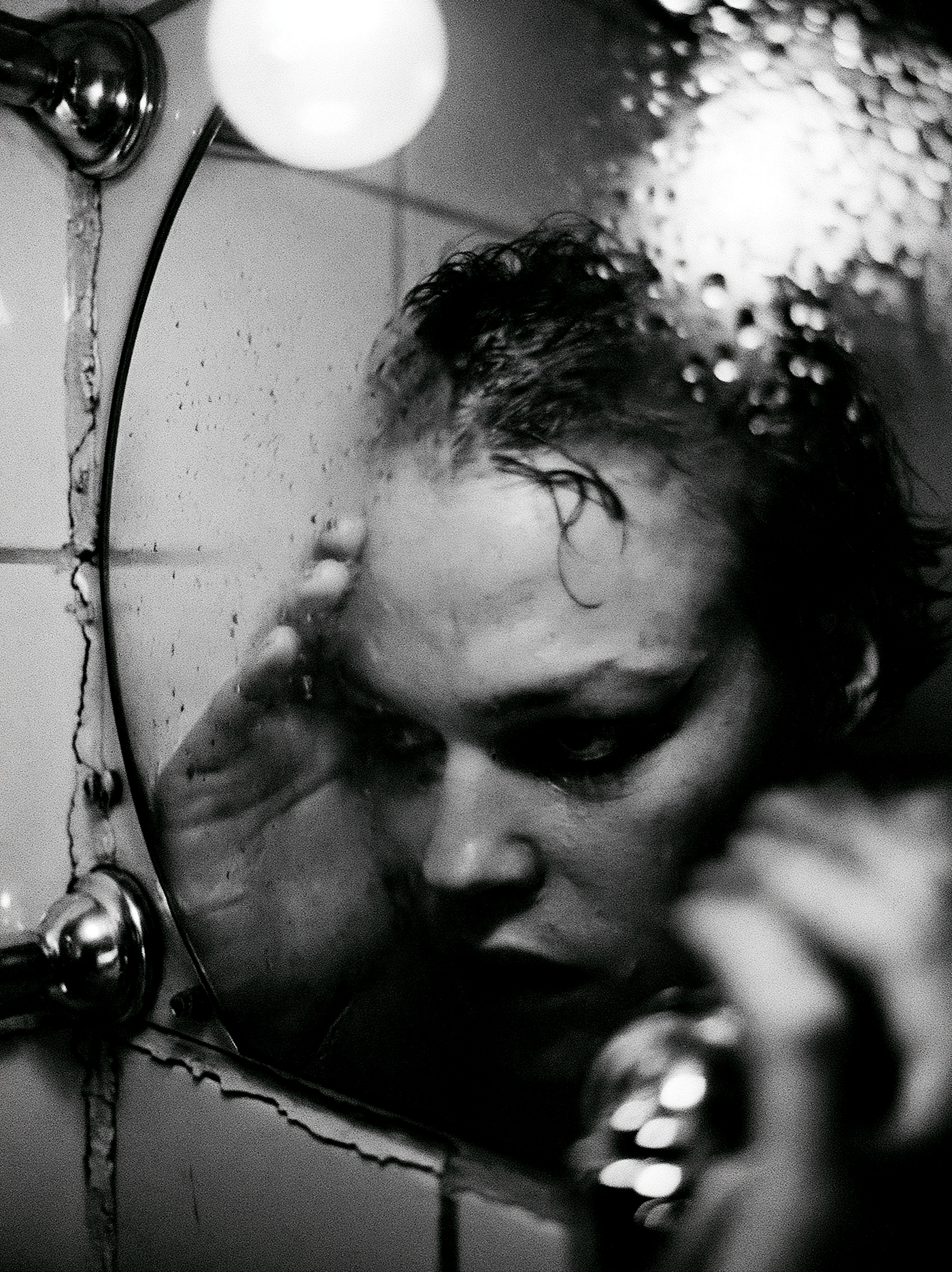 Prompt: medium-close grainy black and white editorial portrait of a 30-year-old Swedish woman with smudged eyeliner and damp short hair, leaning her forehead against a fogged bathroom mirror in a 1970s Stockholm apartment, photographed in the spirit of Anders Petersen, cinematography by Robby Müller, shot on 35mm Ilford HP5 400 pushed to 1600 with Nikon FM2 and 35mm f/1.4 Nikkor, single bare tungsten bulb above the mirror casting hard top-down light with deep eye socket shadows, high contrast monochrome with crushed blacks and blown highlights on the mirror fog, claustrophobic framing with heavy vignetting, cracked tile grout and condensation droplets on chrome fixtures, coarse aggressive grain structure with motion blur on the edges of the hands — ar 3:4 — raw — stylize 80 — v 8.1